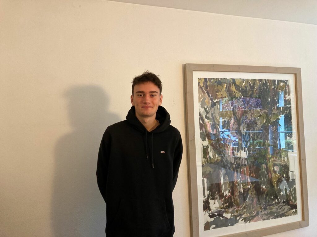 Junger Mann in schwarzem Pullover neben einem Gemälde vor weißer Wand.
