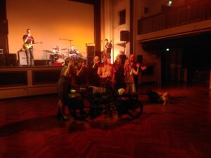 einige Menschen stehen vor einem Tandem-Fahrrad und klatschen in Gebärdensprache (schütteln die Hände), eine Band spielt im Hintergrund auf einer Bühne