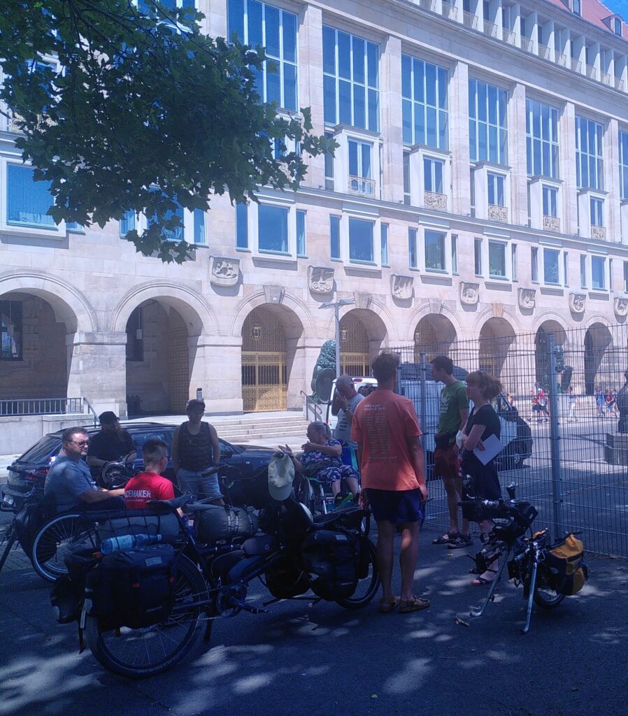 eine Gruppe Menschen steht vor einem Gebäude, es sind Fahrräder und Rollstühle zu sehen