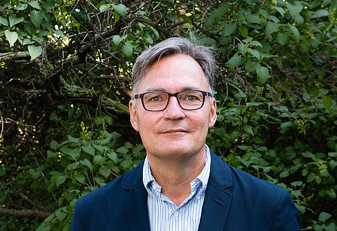 Mann in blauem Anzug mit Brille vor grüner Hecke