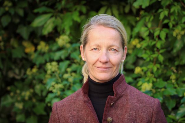Blonde Frau in weinroter Jacke vor grüner Hecke