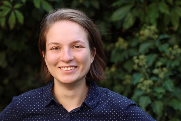 Porträt einer jungen Frau mit braunen Haaren vor einer grünen Hecke