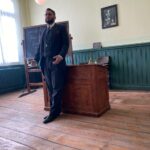 Mann in Kleidung eines Lehrers aus der Kaiserzeit steht am Lehrerpult vor der Klasse