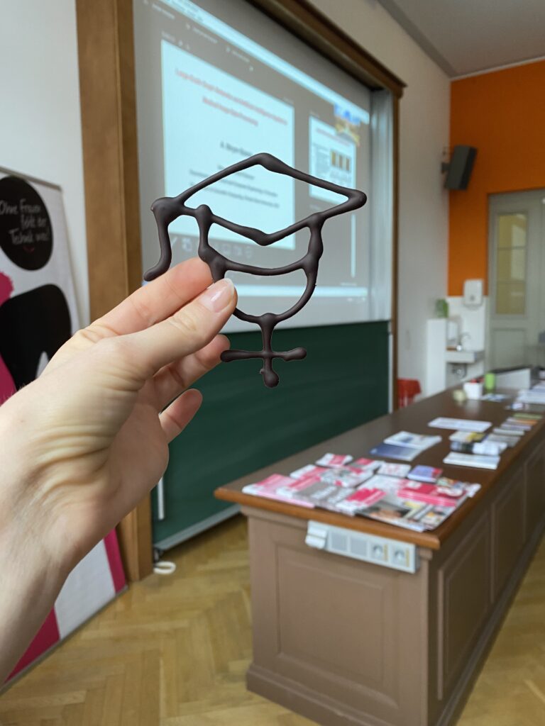 Eine Hand hält eine Form aus Schokolade. Die Form zeigt einen Doktorhut, der auf dem Symbol für das weibliche Geschlecht sitzt. Im Hintergrund befindet sich die Tafel eines Hörsaals und davor ein langes braunes Holzpult, auf dem viele Broschüren liegen.