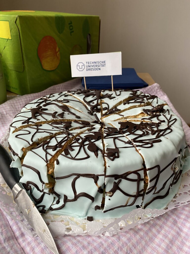 Mit hellblauem Guß überzogene, runde Torte steht auf einem Tisch. Der Guß ist mit Schokoladenspritzern versehen. Die Torte ist in 14 Stücke geschnitten. Darin steckt ein Schild mit der Aufschrift: Technische Universität Dresden. Links daneben das Logo ein U, in das ein T eingelassen ist. Zusammen ergibt es ein Achteck. Im Hintergrund liegen dunkelblaue Servietten und ein grüner Kasten, auf dem sich ein Apfel und die Aufschrift "10 L" befinden.