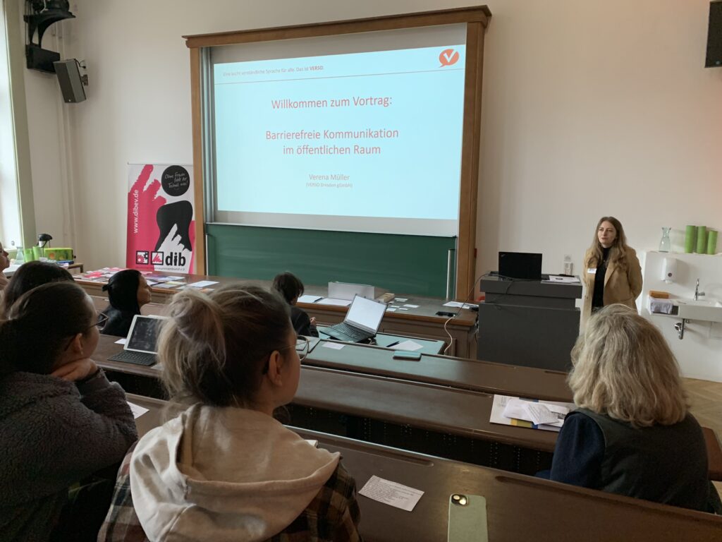 Mehrere Personen sitzen in den Holzbänken eines Hörsaals, Blickrichtung Tafel. Oberhalb der Tafel ist eine Powerpoint-Präsentation an projiziert. Darauf steht in roter Schrift: Willkommen zum Vortrag: Barrierefreie Kommunikation im öffentlichen Raum. Verena Müller (Verso Dresden gGmbH). Am Pult steht eine Person mit langen blonden Haaren in hellbraunem Blazer, darunter ein schwarzer Rollkragenpullover.
