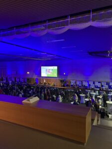 Ein großer Saal aus Perspektive von oben auf der Tribune herab. Im Saal stehen vor einer Bühne viele Stühle auf denen Menschen sitzen. Auf der Bühne befindet sich eine Person. Darüber hängt ein Flachbildschirm, auf dem die Logos der verschiedenen Akteur:innen der Tagung abgebildet sind. Rechts neben den sitzenden Menschen befinden sich mit einer weißen Decke überzogene Stehtische.