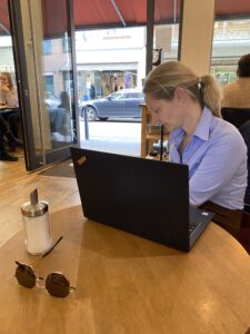 Im Café. Eine blonde Frau mit Zopf in hellblauer Bluse sitzt abgewendet an einem kleinen runden Holztisch und beugt sich seitlich keicht nach unten. Vor ihr steht ein Laptop. Auf dem Tisch steht ein Zuckerstreuer und eine Sonnenbrille liegt darauf. Im Hintergrund befinden sich andere Gäste und eine große geöffnete Eingangstür.