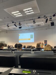 Ein weißer Vortragssaal mit abgestuften Sitzreihen, ähnlich einem Hörsaal, in dem viele Menschen sitzen. Davor eine Bühne, auf der zwei Personen am Pult stehen. An die Wand wird eine Wilkommens-Vortragsfolie projeziert. Darauf befindet sich unter anderem das Logo der diesjährigen Veranstaltung.