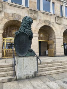 Blick auf ein Gebäude mit großen Goldenen Toren, die in hohe Rundbögen eingelassen sind. Treppenstufen führen zum Gebäude hinauf. Zwischen den Treppenstufen befindet sich die schwarzgrüne Skulptur eines Löwen, der ein Schild mit einer Aufschrift in der Tatze hält.