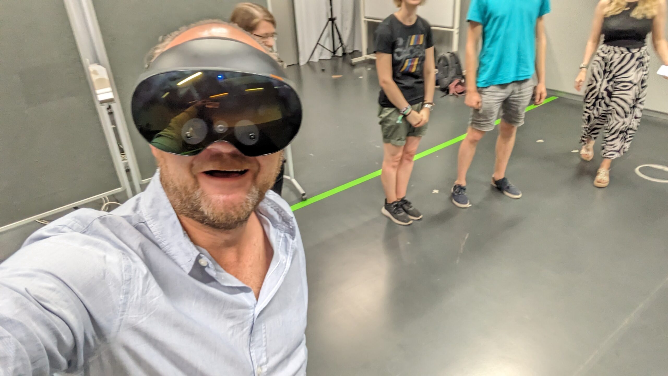 Person mit Halbglatze in hellblauem langärmligen Hemd macht ein Selfie mit schwarzer VR/AR-Brille. Die Brille nimmt fast die Hälfte des Gesichts ein und reicht bis zur Nase. Im Hintergrund befinden sich die Unterkörper vier weiterer Personen. Auf dem dunkelgrauen Boden ist ein neongrüner Streifen, der einen Bereich abteilt.