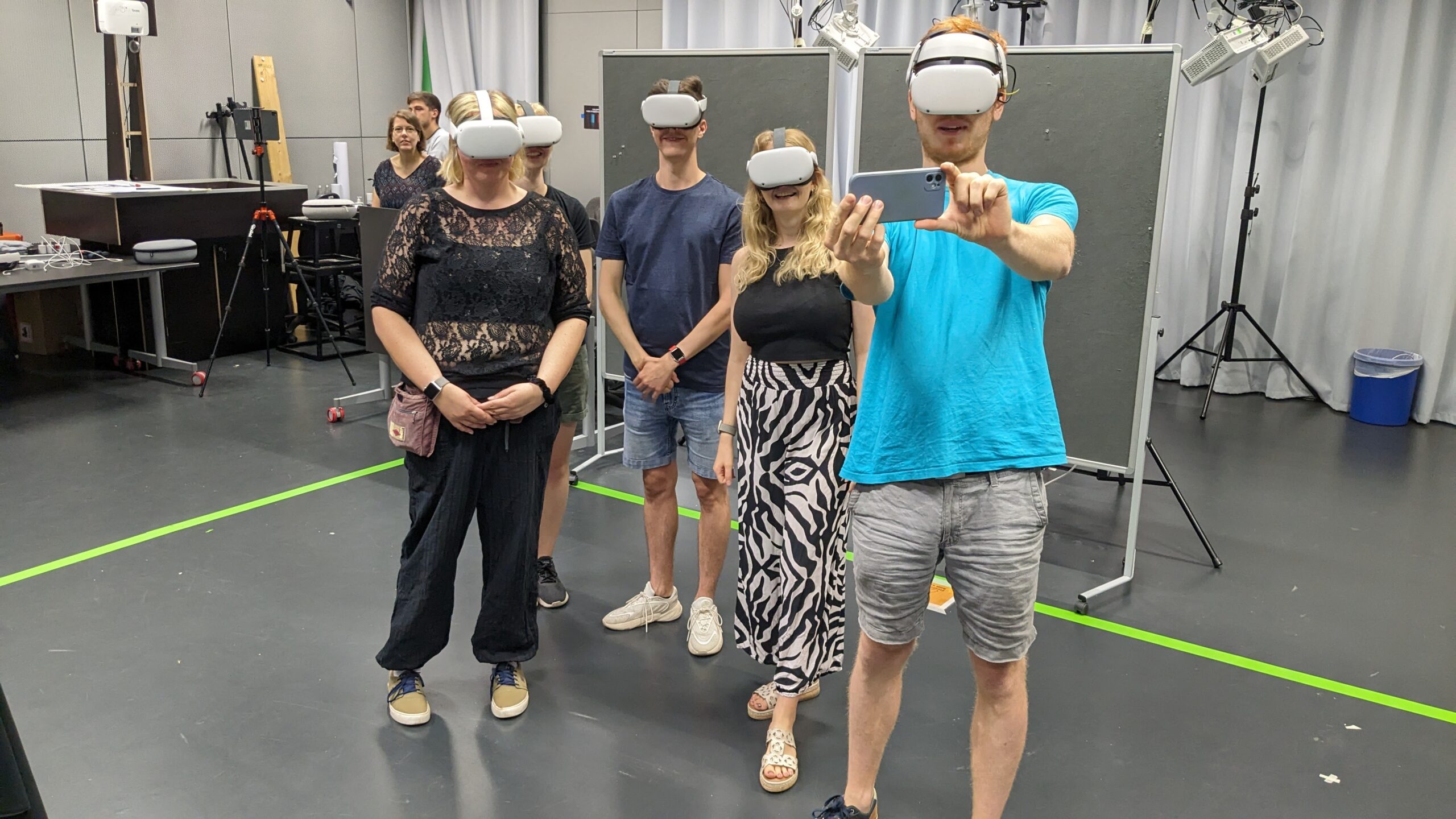 Eine Gruppe von 5 Personen, die dabei fotografiert wurde, während sie ein Selfie macht. Alle tragen graue VR/AR-Brillen, die den Großteil des Gesichts bis über die Nase verdecken. Von links nach rechts: Eine weiblich gelesene Person mit mittellangen blonden Haaren in schwarzem durchsichtigen Spitzenoberteil und schwarzem Crop-Top darunter und schwarzer Hose mit khakifarbenen Sneakern mit dunkelblauen Schnürsenkeln. Dahinter größtenteils verdeckt eine weiblich gelesene Frau mit kurzen blonden Haaren in schwarzem T-Shirt und dunkelgrüner Shorts und grauen Sportschuhen. Daneben eine männlich gelesene Person mit kurzen braunen Haaren in dunkelblauem T-Shirt, Jeans-Shorts und weißen Sneakern. Daneben eine weiblich gelesene Person mit blonden langen haaren, einem schwarzen kurzen Oberteil und eine weiß-schwarz gemusterten weiten Pluderhose und beigefarbenen Sandalen mit Absatz. Daneben eine männlich gelesene Person mit roten Haaren und rotem kurzem Bart in türkisfarbenem T-Shirt und grauer Jeans-Shorts. In den Händen hält die Person ein Smartphone. Im Hintergrund befinden sich sämtliche technische Geräte. Auf dem dunkelgrauen Boden ist eine neongrüne Linie, die einen Bereich abtrennt.