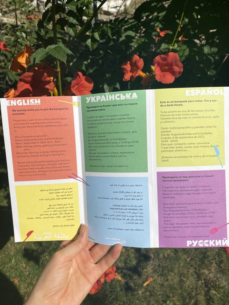 Hand hält aufgeklappten Flyer vor Hecke mit roten Blüten. Auf dem Flyer sind sechs bunte Felder jeweils in unterschiedlichen Sprachen von links nach rechts (oben): Englisch, Ukrainisch, Spanisch und unten: Arabisch, Farsi/Persisch und Russisch.