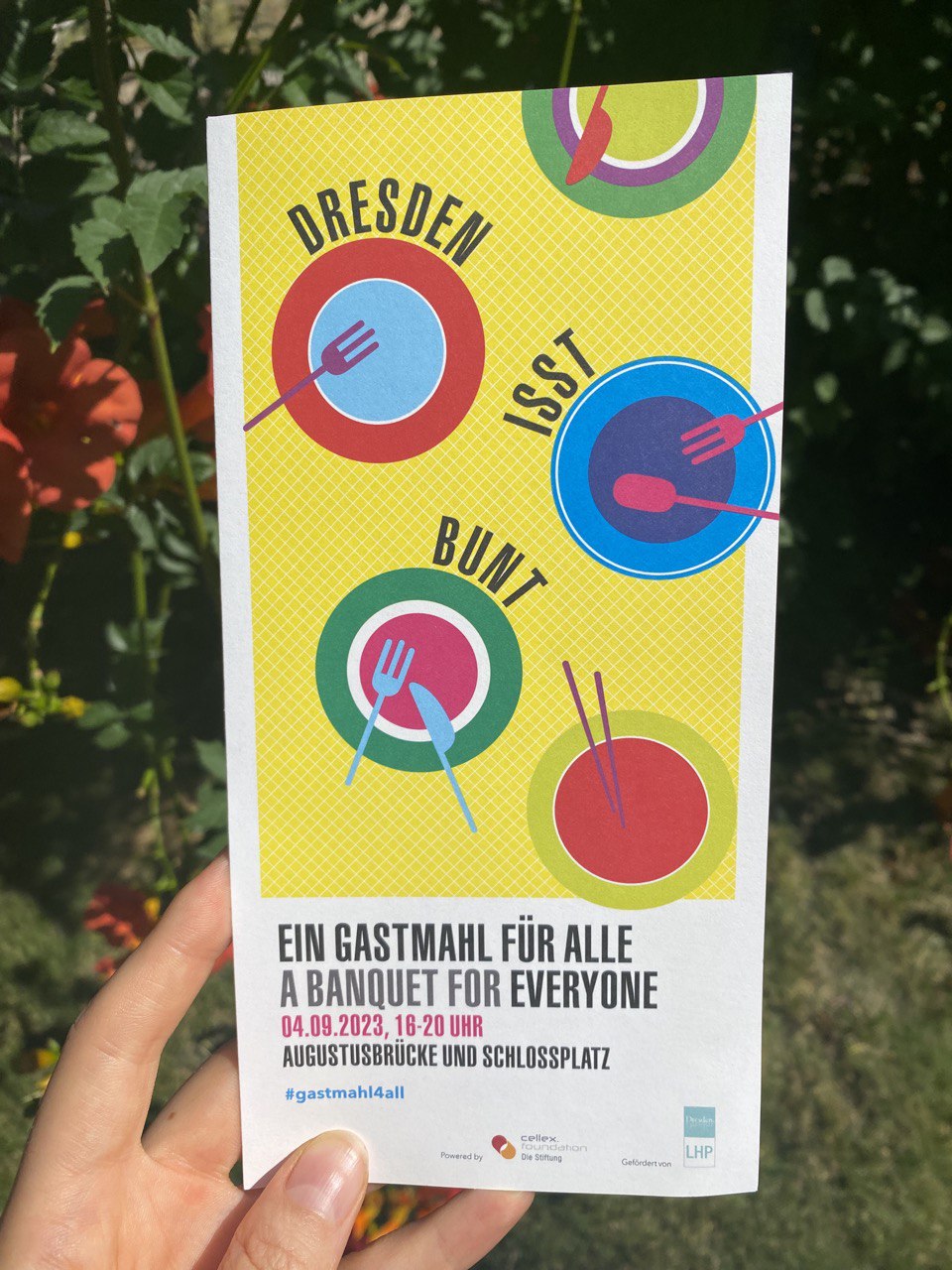 Eine Hand hält einen bedruckten Flyer. Im Hintergrund verschwommen eine Hecke mit roten Blüten. Vor gelben Hintergrund sind auf dem Flyer bunte Teller mit verschiedenem bunten Besteck gedruckt. Darunter steht "Ein Gastmahl für alle. A banquet for everyone. 04.09.2023, 16-20 Uhr. Augustusbrücke und Schlossbrücke. #gastmahl4all. Unten kleine Logos "powered by Cellex foundation die Stiftung, gefördert von LHP Dresden."
