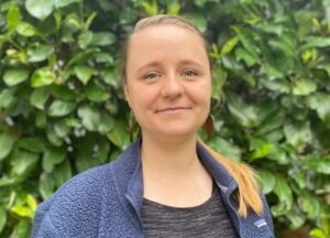Weiblich gelesene Person vor grüner Hecke lächelt, trägt blaue Strickjacke und schwarzes Top, blonde Haare zu Zopf gebunden