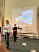 Alexander Lasch vor dem Publikum, dahinter Juliane Heidelberger und die Leinwand