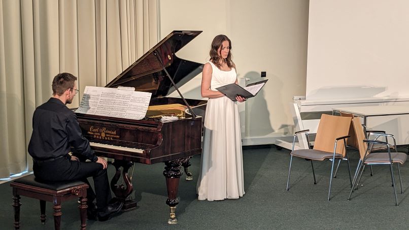 Mann in schwarzem Anzug am Flügel, Frau in weißem Hochzeitskleid steht singend davor