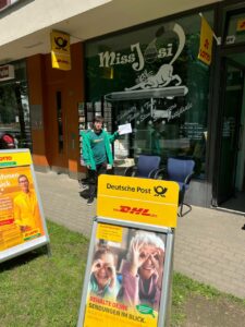 Clemens in grüner Jacke vor dem Eingang einer Postfiliale, er hält den weißen Brief hoch