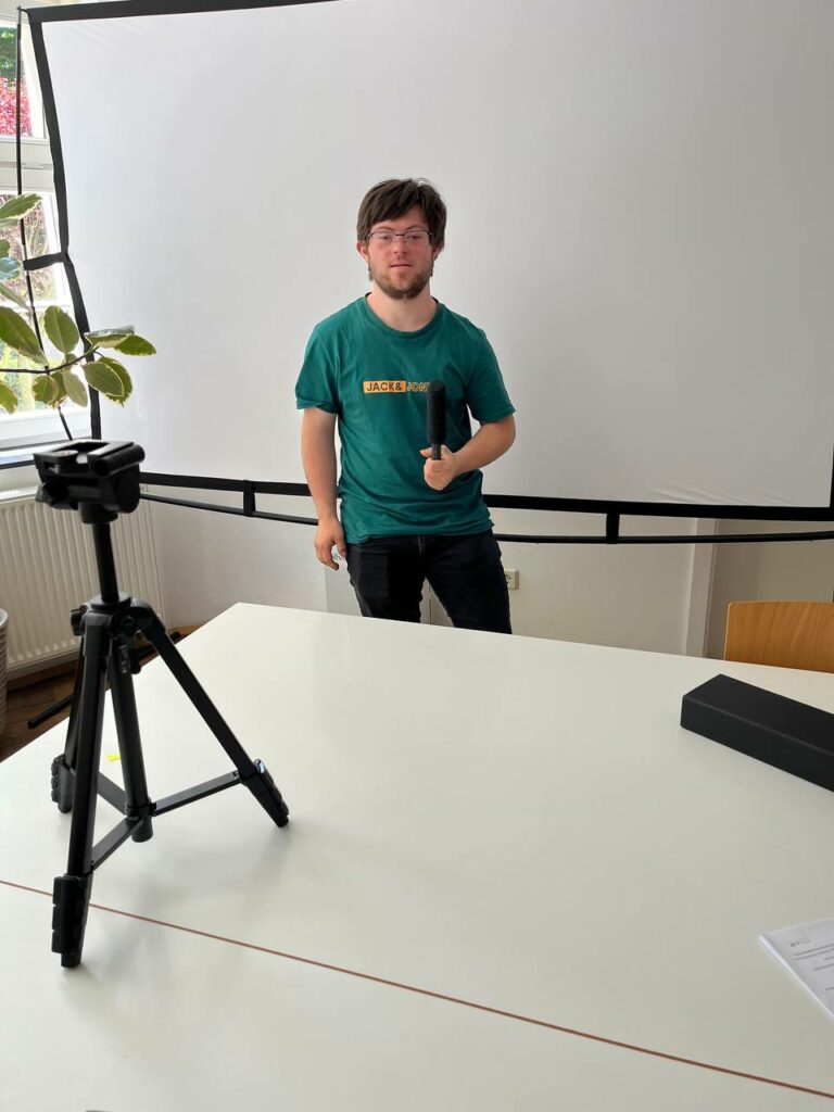 Clemens in grünem T-Shirt vor weißer Leinwand, er zeigt verschiedene Posen mit dem Mikro in der Hand