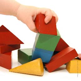 vor weißem Hintergrund bunte Holzbausteine, eine Kinderhand hält davon einen roten Baustein