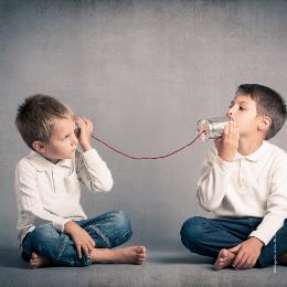 vpr grauem Hintergrund sitzen 2 Jungs in Jeans und weißen Hemden im Schneidersitz auf dem Boden, sie halten sich jeder eine Blechdose ans Ohr, die mit einer roten Schnur verbunden sind