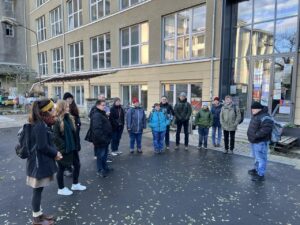 Personen von TTT macht inklusive Schule in Winterkleidung vor dem Eingang eines mehrstöckigen Gebäudes mit großen Fabrikfenstern