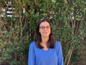weiblich gelesene Person mit offenen schulterlangen braunen Haaren in blauem Pulli mit V-Ausschnitt vor grüner Hecke