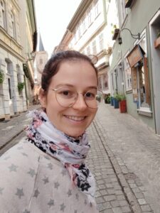 Junge Frau mit braunen Haaren und großer Brille trägt einen hellen Pullover und ein geblümtes Tuch um den Hals, im Hintergrund eine Altstadtgasse