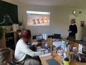 Kursraum mit Leinwand in der Mitte, mehrere Personen von Netz Bangladesch sitzen um einen Konferenztisch und schauen nach vorn, Juliane steht rechts und eröffnet den Kurs