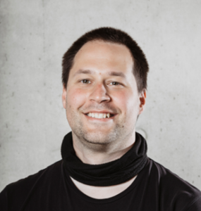 Lächelnde männlich gelesene Person mit kurzen braunen Haaren, schwarzem Pullover und schwarzem Tuch um den Hals